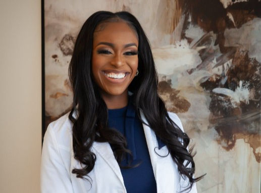 Smiling woman in a white coat stands in front of abstract art. Her dark hair contrasts with the light and dark tones of the background.
