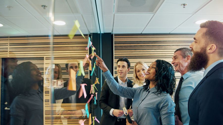 The photo shows a diverse group of professionals gathered around a glass wall with colourful sticky notes, where a woman is leading the discussion with a smile.
