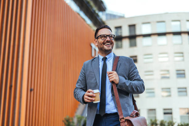 Confident businessman with glasses and a beard enjoys a coffee while walking outside an office building in the city.