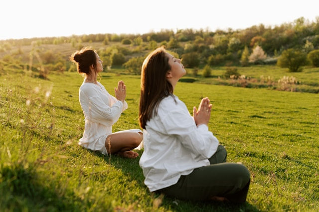 Two women meditate on a grassy hillside at sunset, hands together, facing the warm, green countryside.