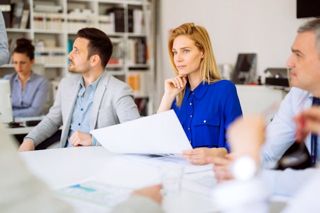 People in a modern office setting engaged in a meeting. A woman in a blue shirt holds papers, looking thoughtful. Bookshelves in the background.