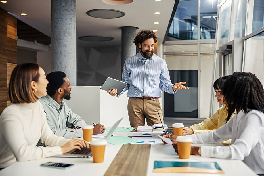 Man presents with tablet in hand to a group around a table with laptops, papers, and coffee cups in a modern office. Collaborative mood.