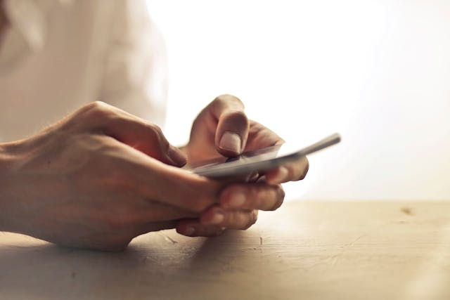 Hands holding a smartphone, typing with focus. Soft lighting, wooden surface, and blurred white background create a calm mood.