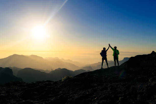 Two silhouetted hikers high-five on a mountain ridge at sunrise, with hazy peaks and a blue-gold sky.