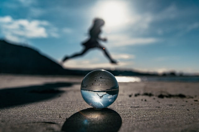 Clear glass ball on sandy beach with blurry person jumping in the sunny background. Bright sky and ocean visible. Energetic mood.