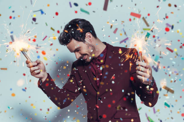 A man in a maroon suit holding sparklers in both hands, surrounded by colorful confetti, celebrating joyfully.