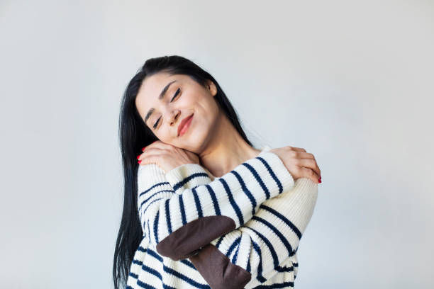 Woman with closed eyes hugging herself, wearing a striped sweater with elbow patches. Gray background, serene and content mood.