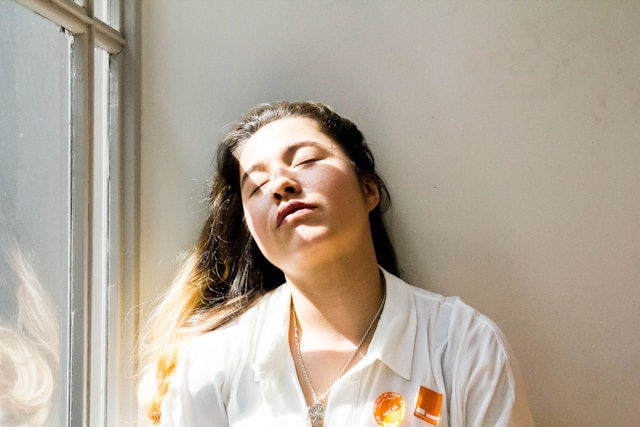 Sunlit woman in a white shirt leans against a wall by a window, eyes closed in a calm, relaxed pose.