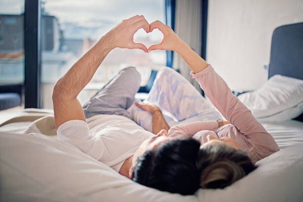 A couple is lying on a bed together, forming a heart shape with their hands above them.