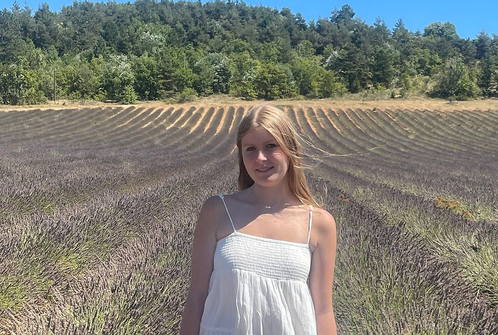 Woman in white dress smiling in a lavender field under blue sky. Rows of lavender stretch into the distance, bordered by green trees.