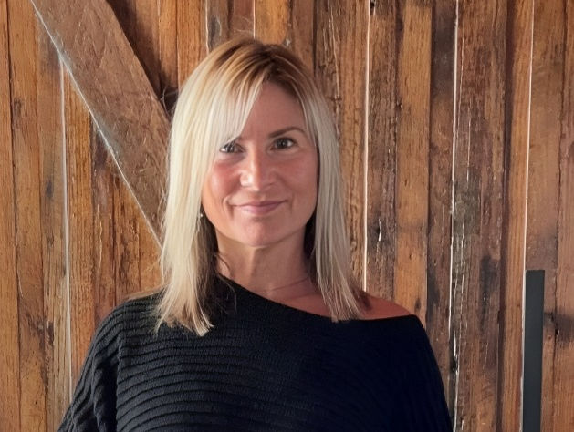 Smiling blonde woman in a black top stands before a rustic wooden wall, posing calmly indoors.