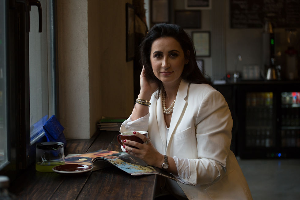Woman in white blazer holding a cup, sitting by a window in a cafe. A magazine is open on the table. Dim lighting with a relaxed mood.