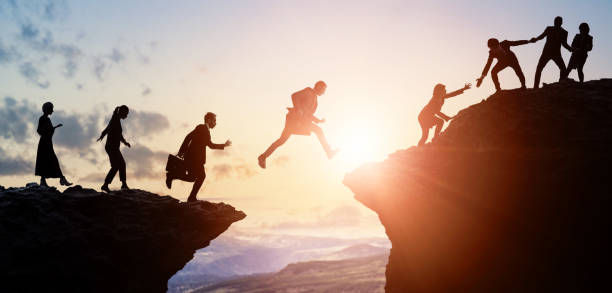 Silhouettes of people helping each other cross a cliff gap at sunrise, symbolizing teamwork and support.