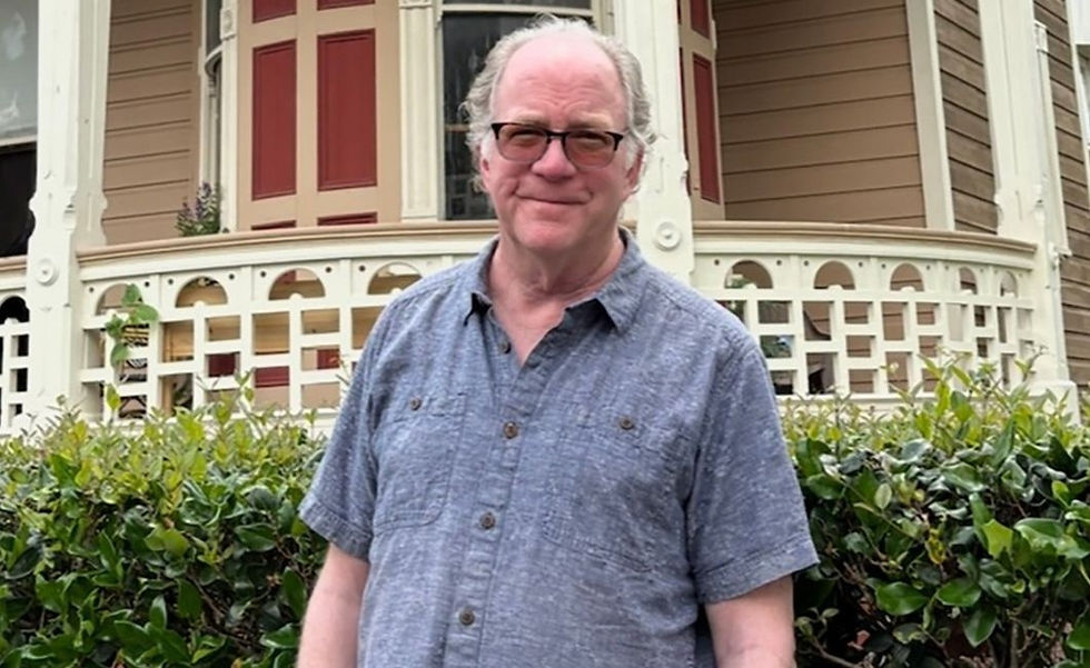 Man in a gray shirt stands smiling in front of a Victorian house with red details and white trim, surrounded by green hedges. Relaxed mood.