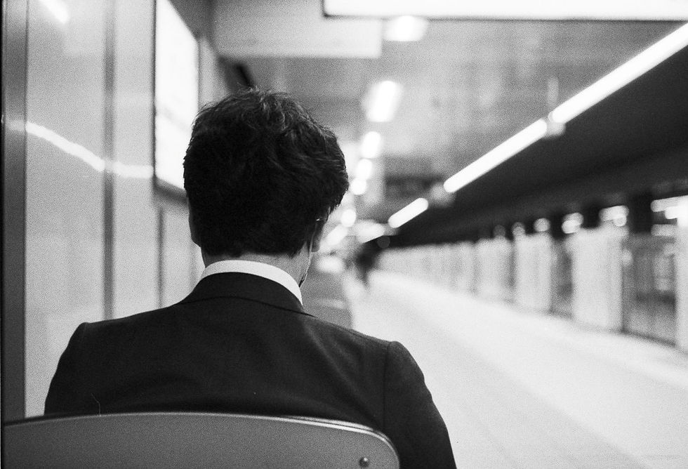 Person in a suit sits on a chair facing a blurred subway platform. Black and white image with a calm, waiting mood.