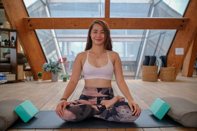 Smiling woman sits cross-legged on a yoga mat in a bright wooden studio, with bolsters and blocks beside her.