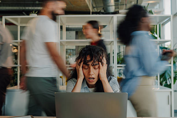 Stressed woman at a laptop in a busy office, holding her head as coworkers blur past in the background.