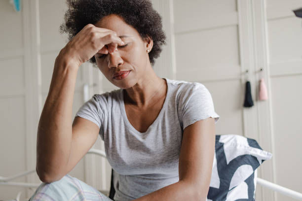 A woman sits on a bed with her eyes closed and her hand on her forehead, appearing stressed or fatigued.