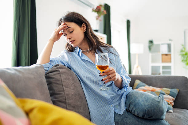 Stressed woman sits on a couch in a bright living room, holding a wine glass and rubbing her forehead.