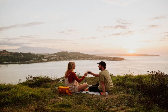 A couple is sitting on a grassy hill overlooking the ocean at sunset, enjoying a picnic and raising glasses in a toast.