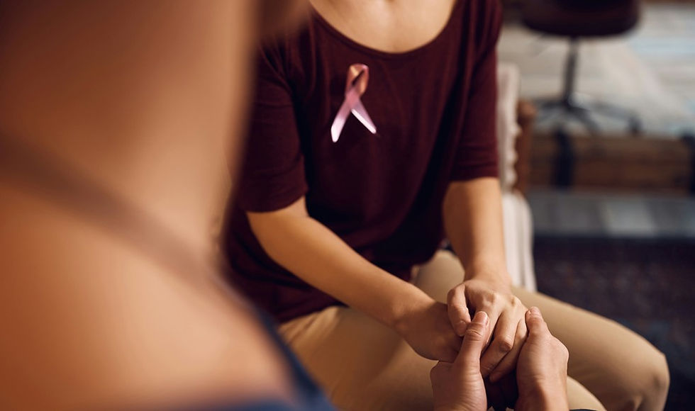 Woman with pink breast cancer ribbon holds another person’s hands in a supportive, intimate counseling setting.