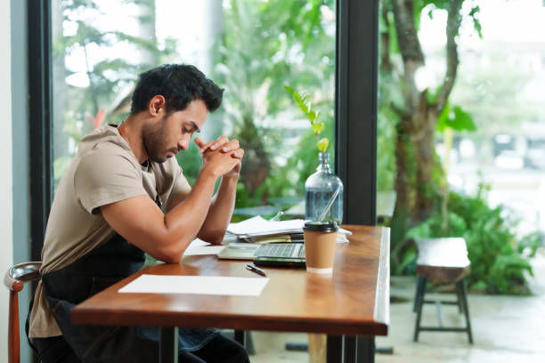 A man sits at a wooden table in a café or office, looking stressed and deep in thought, surrounded by papers, a laptop, and a coffee cup.