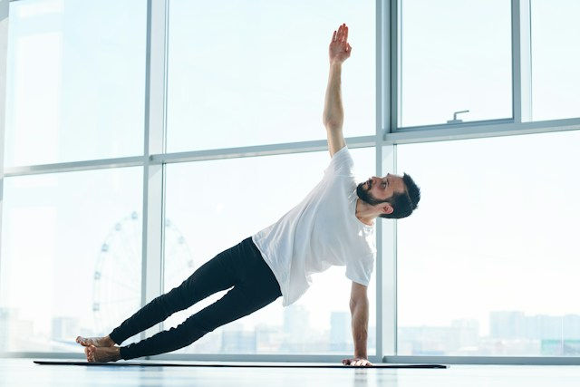 Man doing a side plank yoga pose on a mat in a bright studio with large windows and a city view behind him.