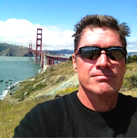 Man in sunglasses takes a selfie with the Golden Gate Bridge and bay in the background. Clear sky and grassy hills create a relaxed mood.