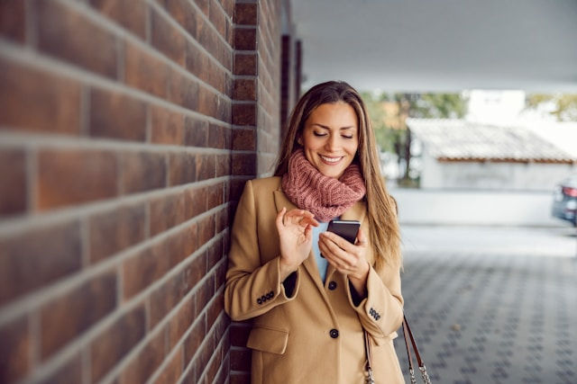 Woman in a tan coat smiles at phone, leaning against a brick wall. She's outside on a tiled walkway, wearing a pink scarf. Casual, happy mood.