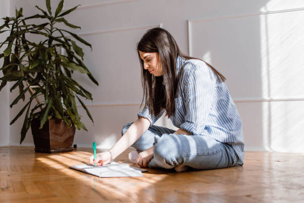 Woman sitting on wooden floor, writing in a notebook with a green pen. Sunlight and plant in background. Casual, focused atmosphere.