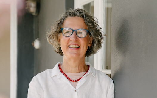 A smiling woman with gray hair and blue glasses stands against a gray wall, wearing a white shirt and red necklace, in a bright setting.