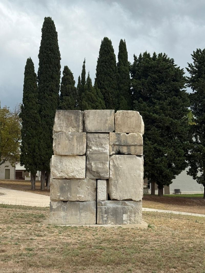 Stacked stone monument in a grassy park, framed by tall cypress trees under a cloudy sky.