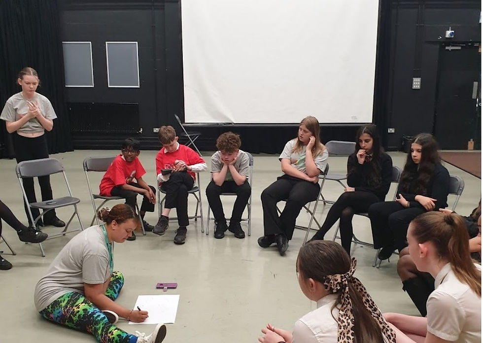 Four people in a counseling circle, one woman taking notes and gesturing, in a bright room with whiteboard and bookshelves.
