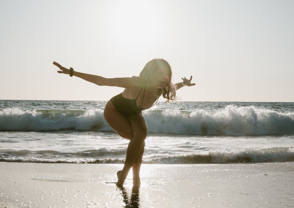 Woman in a black swimsuit playfully poses on a sunny beach with crashing waves in the background, creating a joyful and carefree mood.