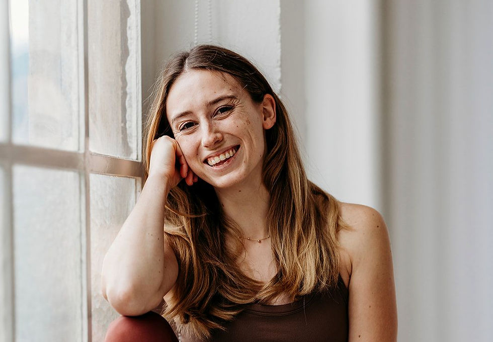 Smiling young woman with long brown hair leans on her hand by a sunlit window in a bright room.