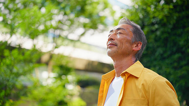 Smiling man in a yellow shirt stands outdoors with eyes closed, basking in bright sunlight amid green trees.