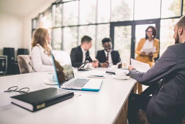 Business meeting in a bright office, four people seated at a table with laptops and documents; one person standing and presenting.