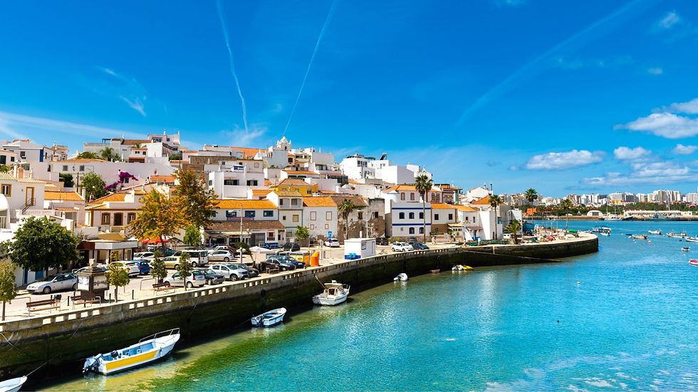 Sunny coastal village with white buildings and orange roofs, overlooking a calm blue river. Moored boats line the waterfront under a clear sky.