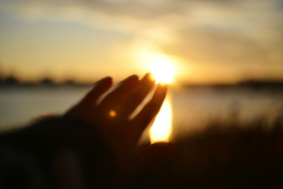A hand reaches toward a bright sunset over a blurred water scene, creating a warm and serene atmosphere.