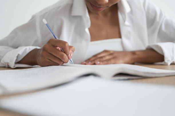 Person writing in a notebook with a pencil, wearing a white shirt. Focused expression in a bright, simple room. Pages are blurred in the foreground.