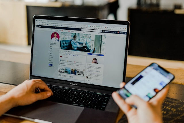 The photo shows a person using a MacBook Pro with Facebook open on the screen while holding a smartphone in their other hand.