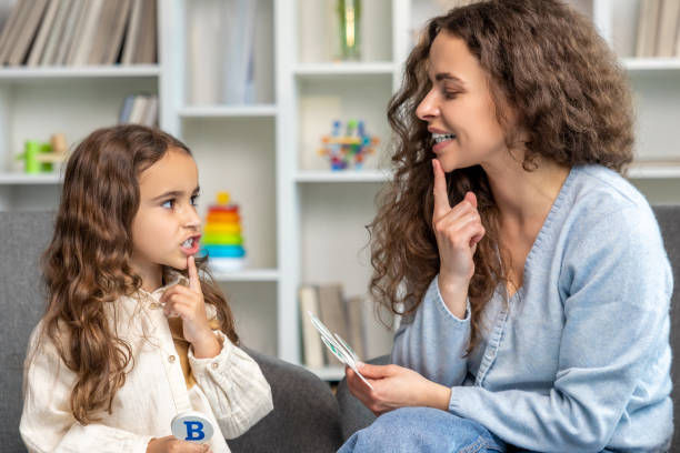 Woman and girl practicing speech therapy in a cozy room. Both point at their mouths, and the girl holds a card with "B". Shelves in background.