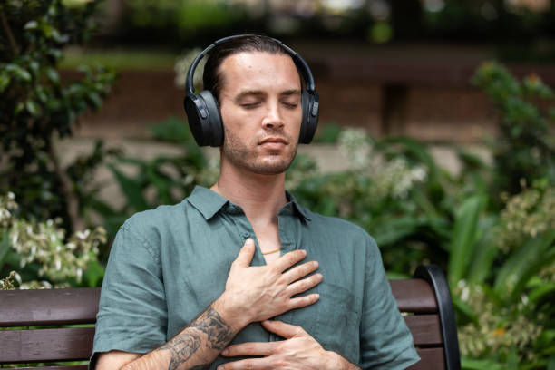 Man with headphones meditating on a park bench, eyes closed, hand on chest. Green shirt, serene expression, lush foliage backdrop.