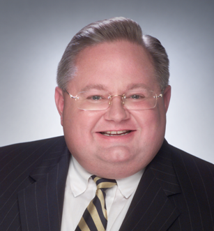 Smiling man in a dark pinstripe suit and striped tie poses against a light gray studio background.