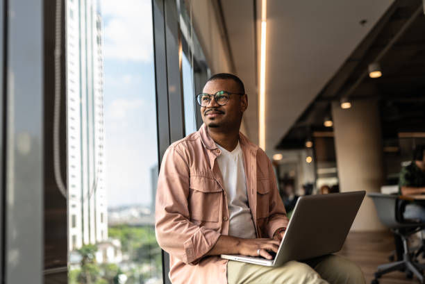 Man in glasses smiles while sitting with a laptop by a large window in a modern office. Cityscape view outside. Casual attire.