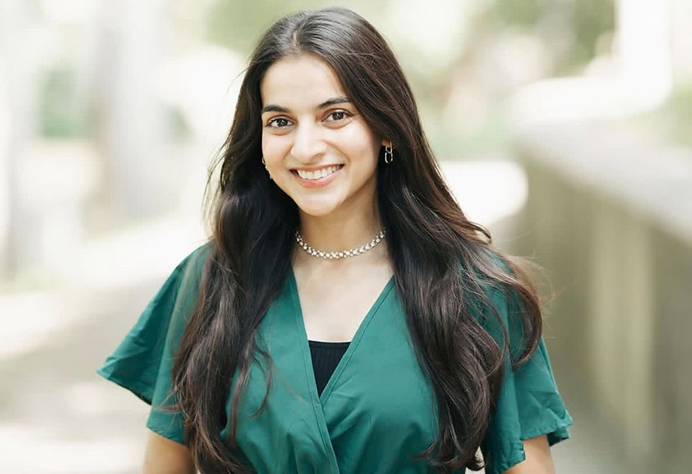 Smiling woman with long hair in a green dress and necklace, outdoors with blurred trees in the background, exuding a cheerful mood.