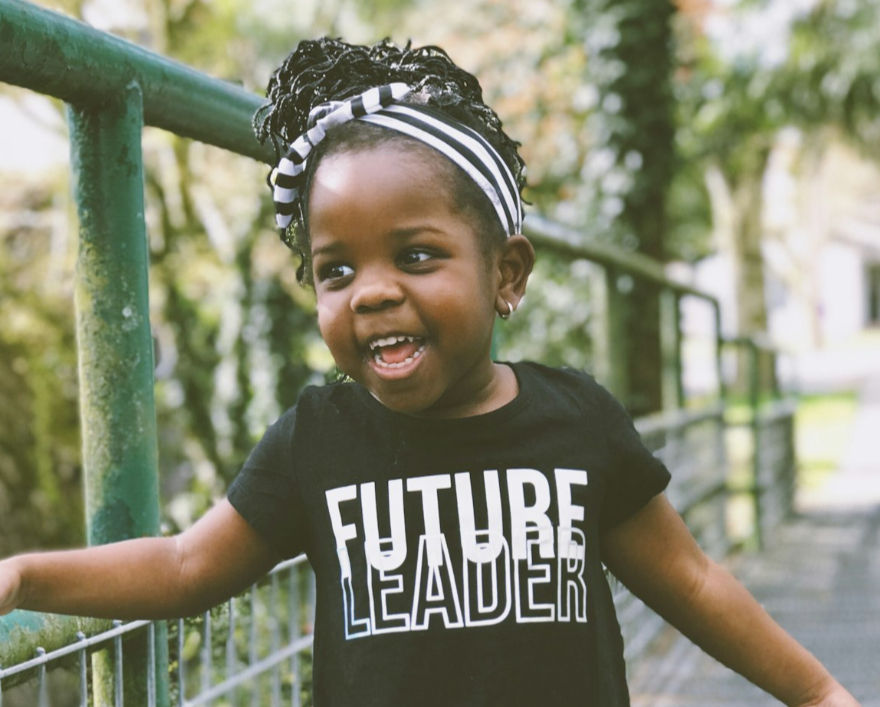 Smiling child in a "Future Leader" t-shirt and striped headband, standing outdoors near a green railing with blurred greenery behind.