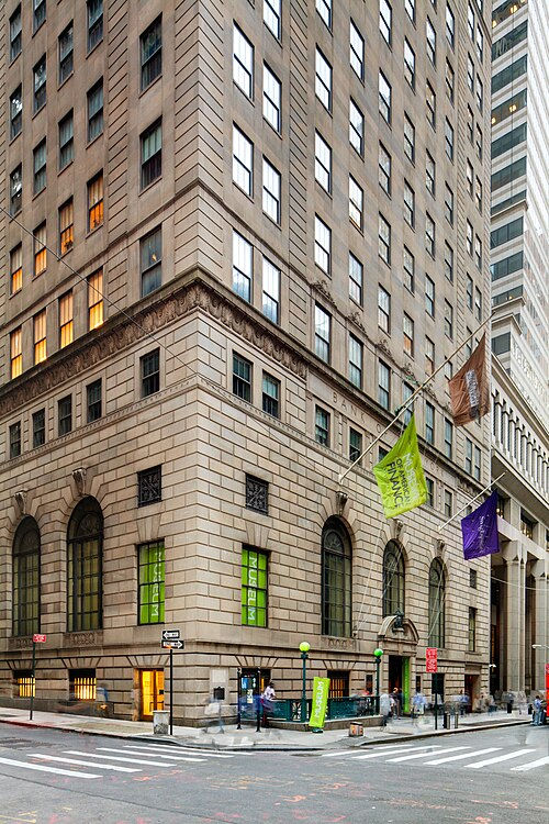 Historic urban building with stone facade, two green flags, a purple flag, and visible street signs. Pedestrians walk by on a city street.