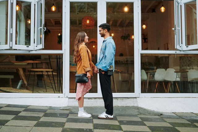 A couple faces each other, holding hands outside a café with large windows. They appear happy. She's in a tan coat, he's in a blue jacket.