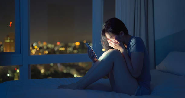 Young woman sits on a bed by a night window, covering her face while looking at a phone, lit by blue city glow.